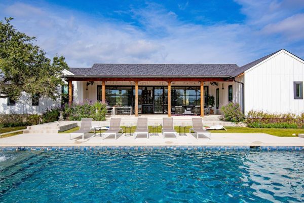 A wide shot of a modern white farmhouse featuring a large swimming pool, several lounge chairs, and a covered patio, a premier Walker Luxury estate.
